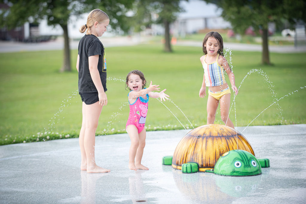 Children playing in a splash pad, laughing and running through water sprays around a turtle-shaped feature in a park.